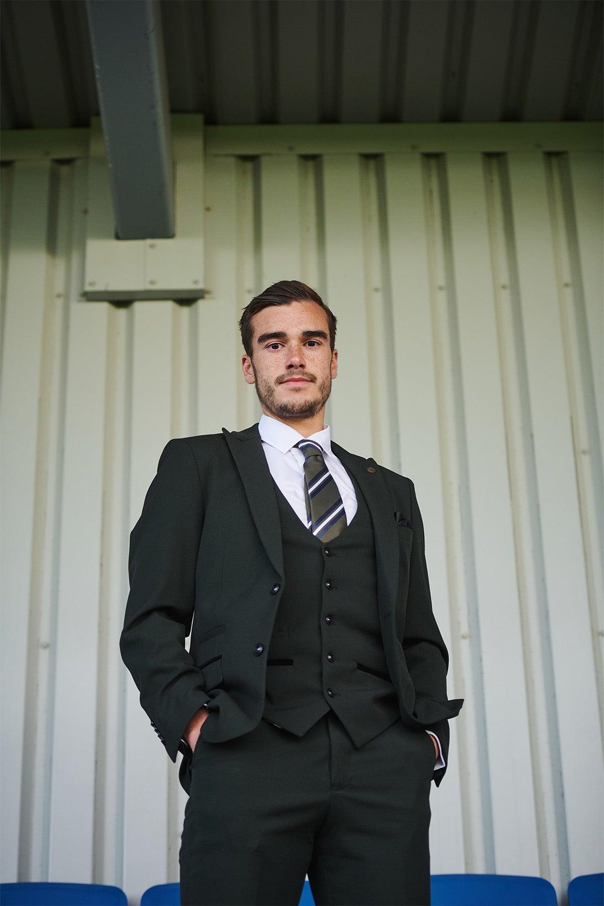 A man in the Marc Darcy Leicester City F.C. Collection BROMLEY Olive Green Check Suit, as worn by Harry Winks, stands with hands in his pockets against a metal-paneled wall, looking confidently at the camera.
