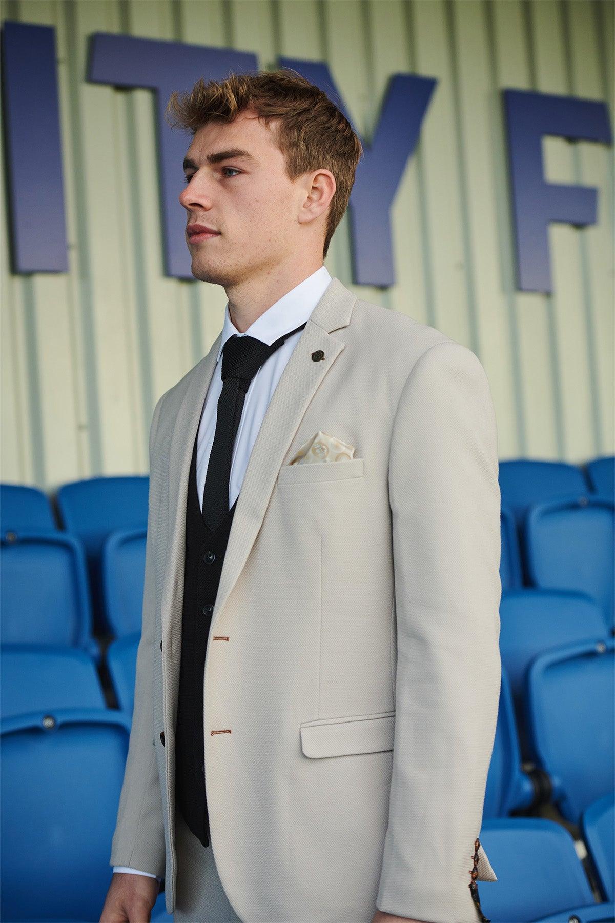 Mads Hermansen wears the Marc Darcy Leicester City F.C. Collection HM5 Stone Suit with Kelvin Black Waistcoat, standing before blue stadium seats, neutral-faced, large wall letters behind him.