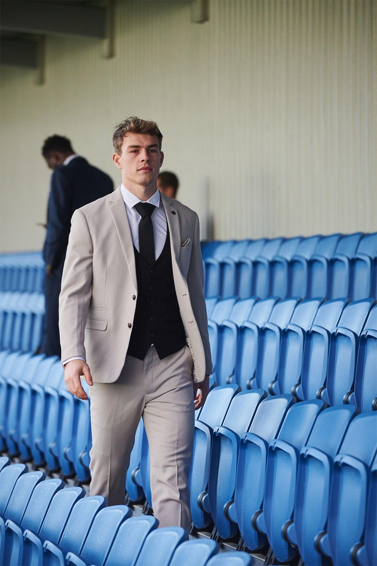 A young man wears the Marc Darcy Leicester City F.C. Collection HM5 Stone Suit with Kelvin Black Waistcoat, as worn by Mads Hermansen, walking through empty blue stadium seats. Another person in a dark suit stands in the background.