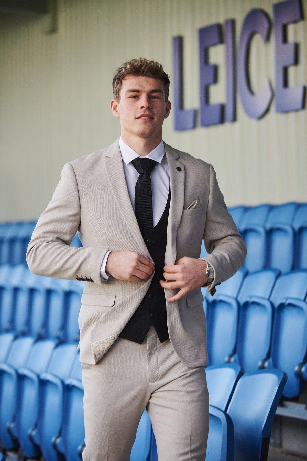 A young man wears Marc Darcy’s Leicester City F.C. Collection HM5 Stone Suit with Kelvin Black Waistcoat, as worn by Mads Hermansen, adjusting his jacket in front of blue stadium seats and a wall displaying "LEICE.