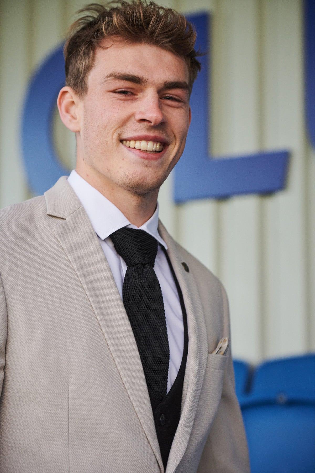 A young man sports the Marc Darcy Leicester City F.C. HM5 Stone Suit with Kelvin Black Waistcoat, as worn by Mads Hermansen, smiling confidently before a blurred backdrop featuring blue letters.