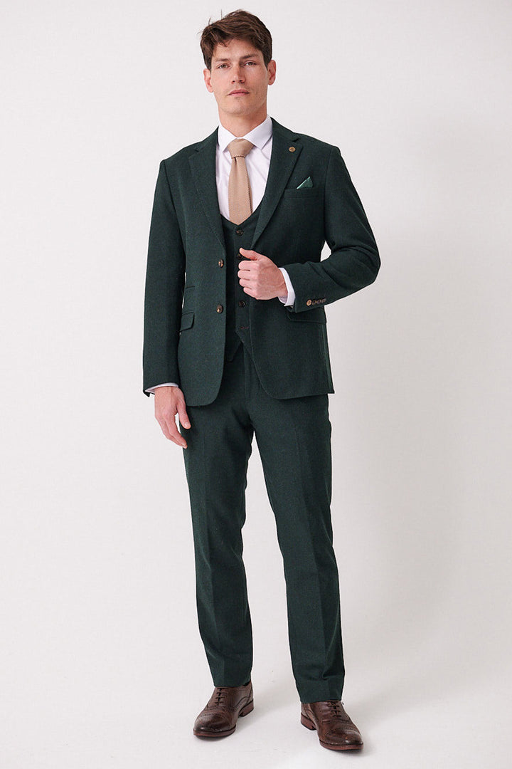 A man with short brown hair and a neutral expression wears the marcdarcy ARCHIE - Forest Tweed Three Piece Suit, featuring a slim fit jacket, matching vest, and trousers, paired with a white shirt, tan tie, and brown dress shoes against a white background.