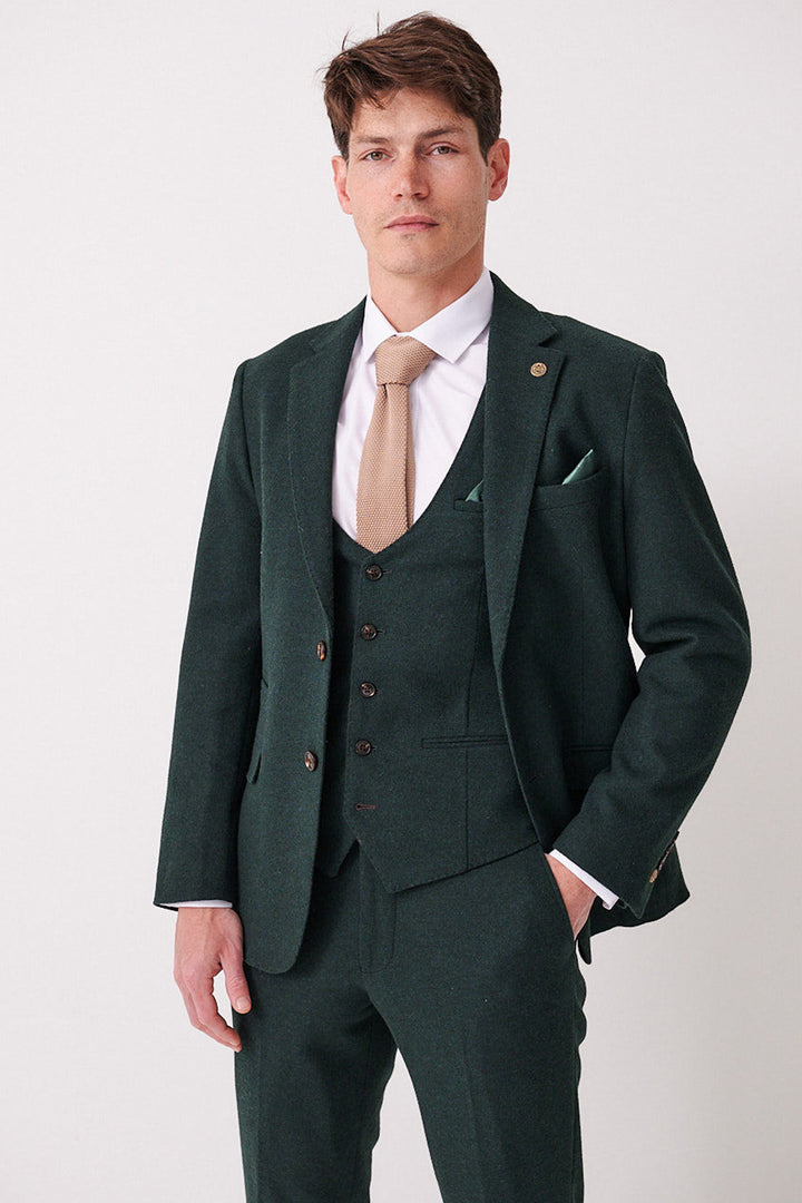 A man with short hair and a neutral expression stands against a white background, wearing the marcdarcy ARCHIE - Forest Tweed Blazer with a matching vest, white dress shirt, light brown tie, and one hand in his pocket.