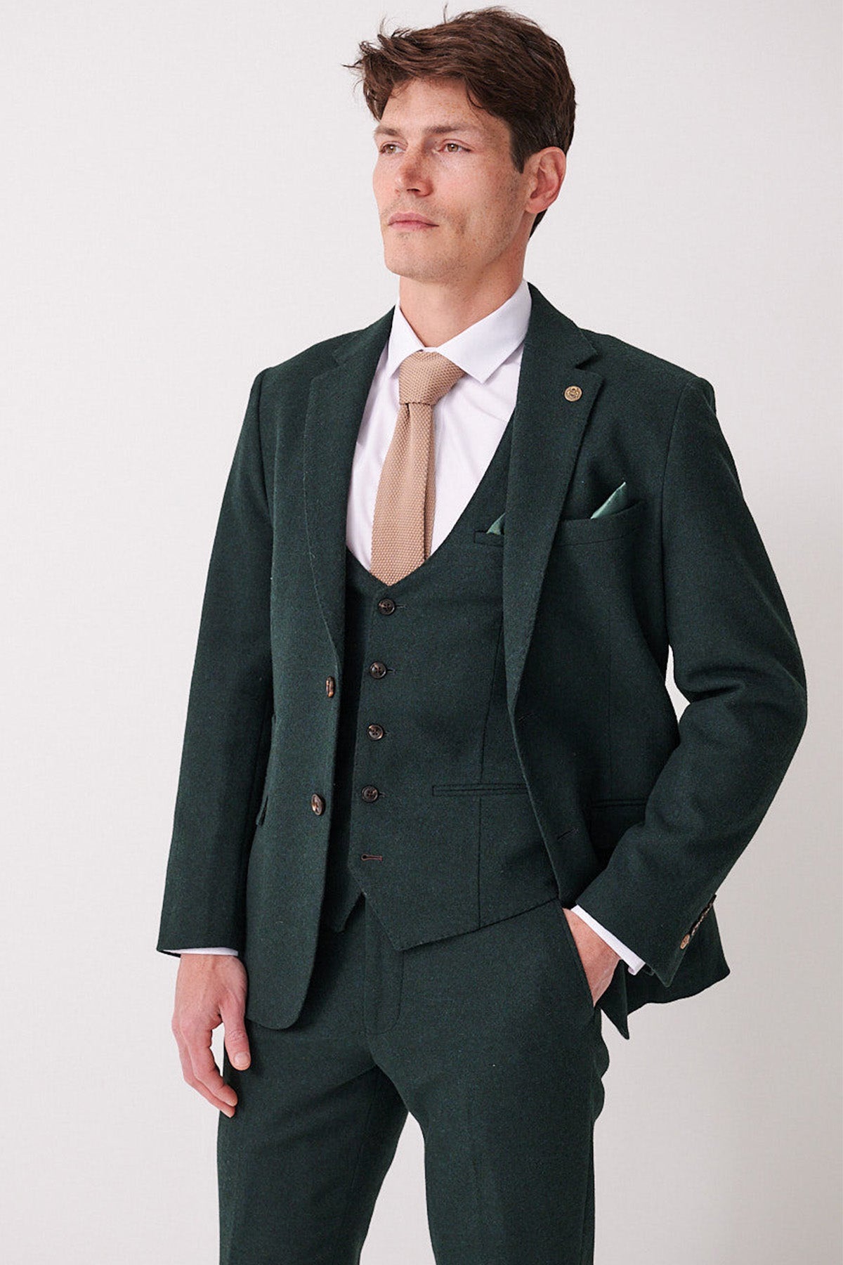 A man models the marcdarcy ARCHIE Forest Tweed Three Piece Suit with a white shirt and tan tie, standing against a plain light background and looking to the side with his left hand in his pocket.