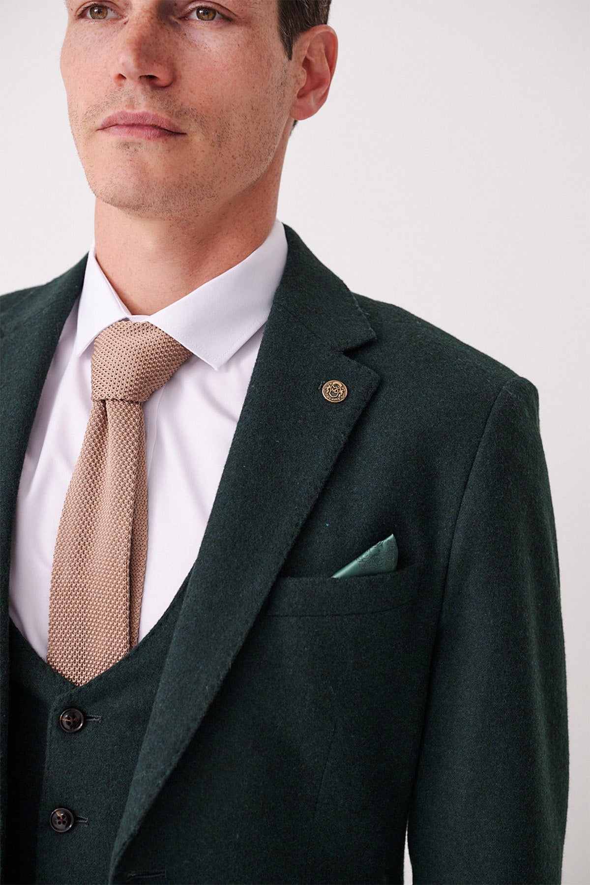 A man wears the marcdarcy ARCHIE - Forest Tweed Blazer with a white dress shirt, beige textured tie, and a green pocket square. Only his lower face and upper torso are visible.