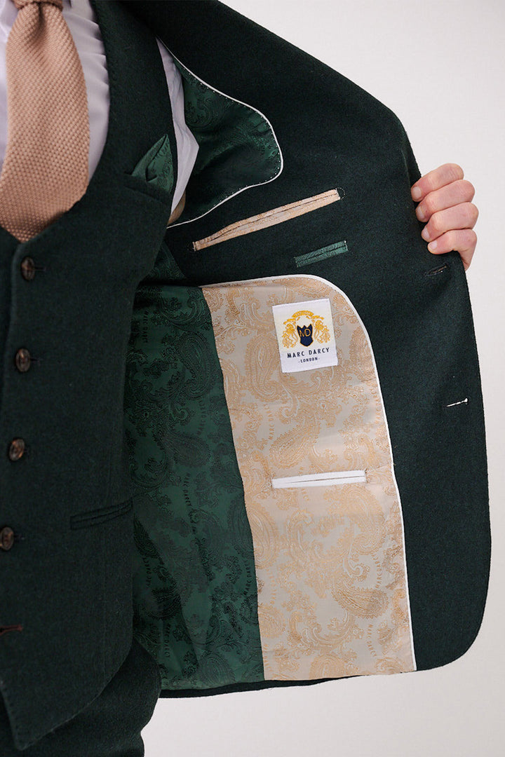 A man in a dark green ARCHIE - Forest Tweed Three Piece Suit by marcdarcy opens his jacket to reveal a paisley green and gold lining, branded label, and multiple inner pockets, highlighting the suit’s elegant slim fit design.