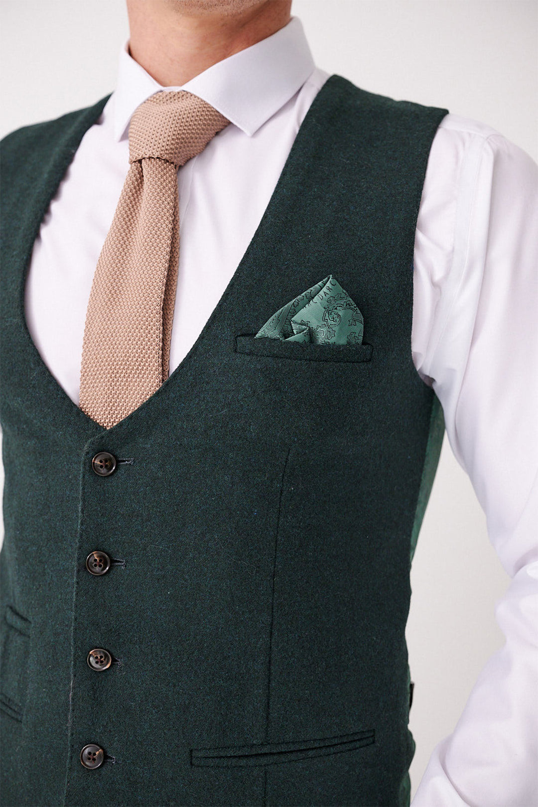 A person wears the marcdarcy ARCHIE - Forest Tweed Three Piece Suit, featuring a dark green vest with a green pocket square, paired with a slim fit white shirt and a light brown textured tie, shown from shoulders to waist.