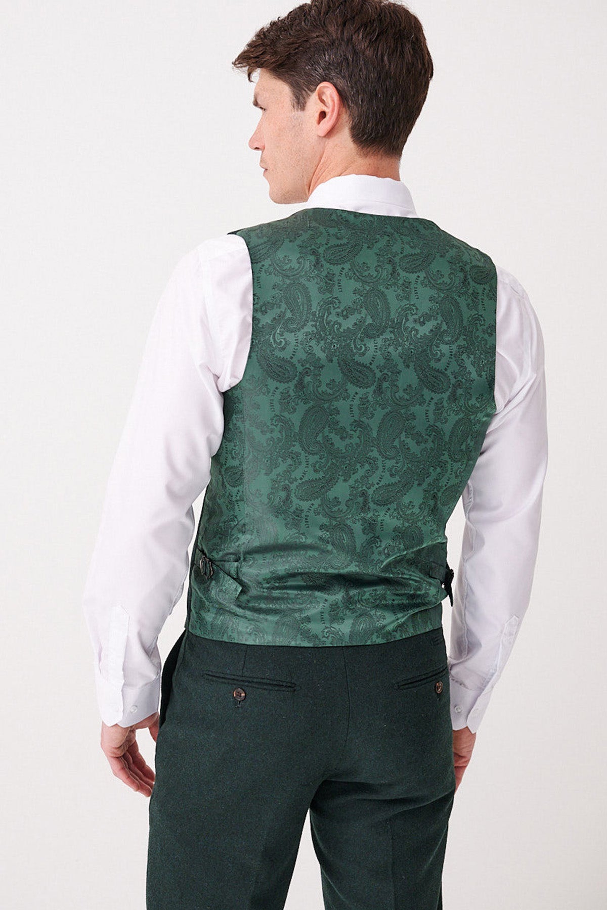 A man facing away wears a white dress shirt, dark green pants, and the Marc Darcy ARCHIE - Forest Tweed Waistcoat featuring an intricate green paisley pattern on the back.