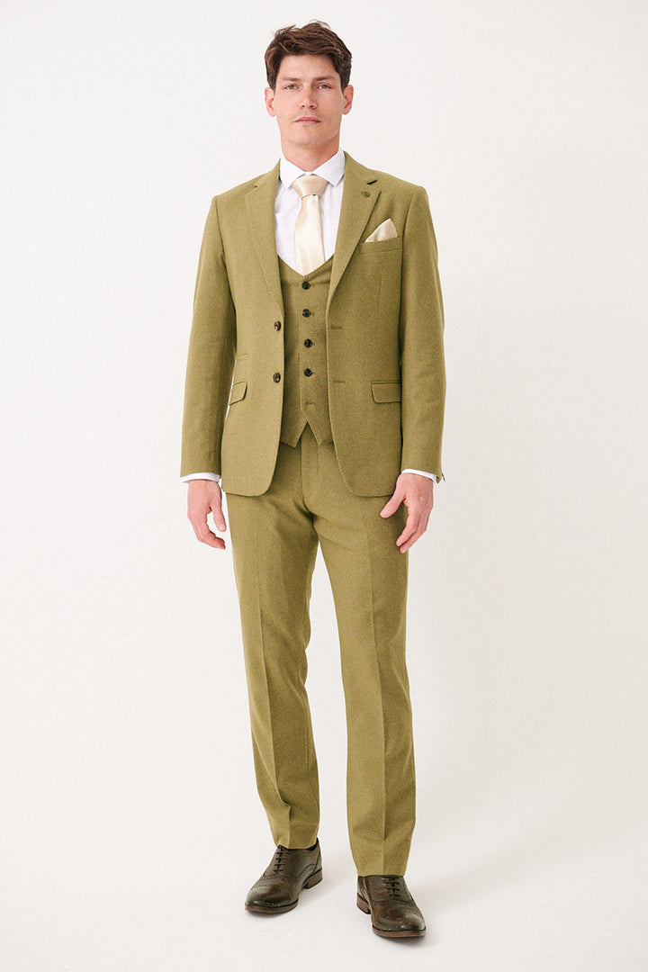 A man faces forward wearing the marcdarcy ARCHIE Moss Tweed Three Piece Suit with matching vest, white shirt, cream tie, and pocket square. He has short brown hair, dark dress shoes, and stands against a plain light background.