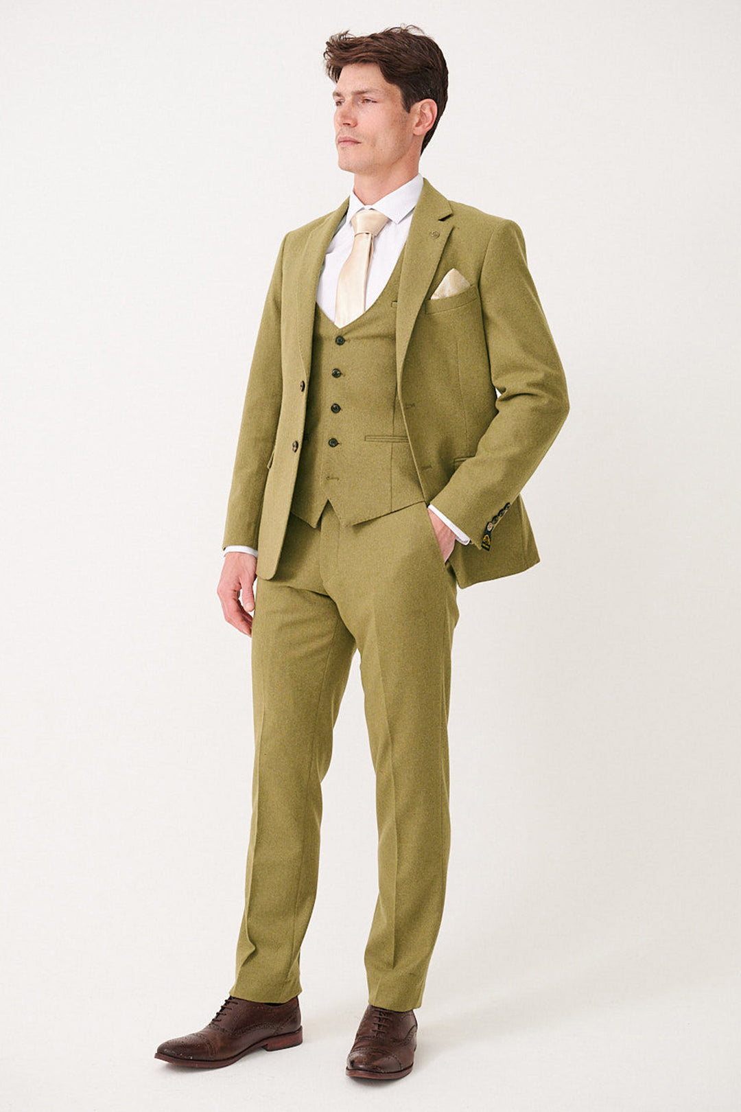 A man wears the marcdarcy ARCHIE Moss Tweed Three Piece Suit with a white shirt, light gold tie, cream pocket square, and brown shoes. He stands against a plain background, looking to the side with one hand in his pocket.