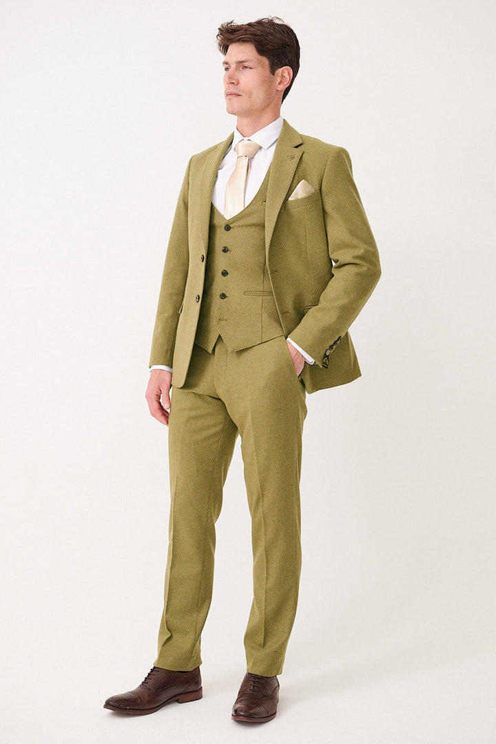A man wears the marcdarcy ARCHIE Moss Tweed Three Piece Suit with a white shirt, light gold tie, cream pocket square, and brown shoes. He stands against a plain background, looking to the side with one hand in his pocket.