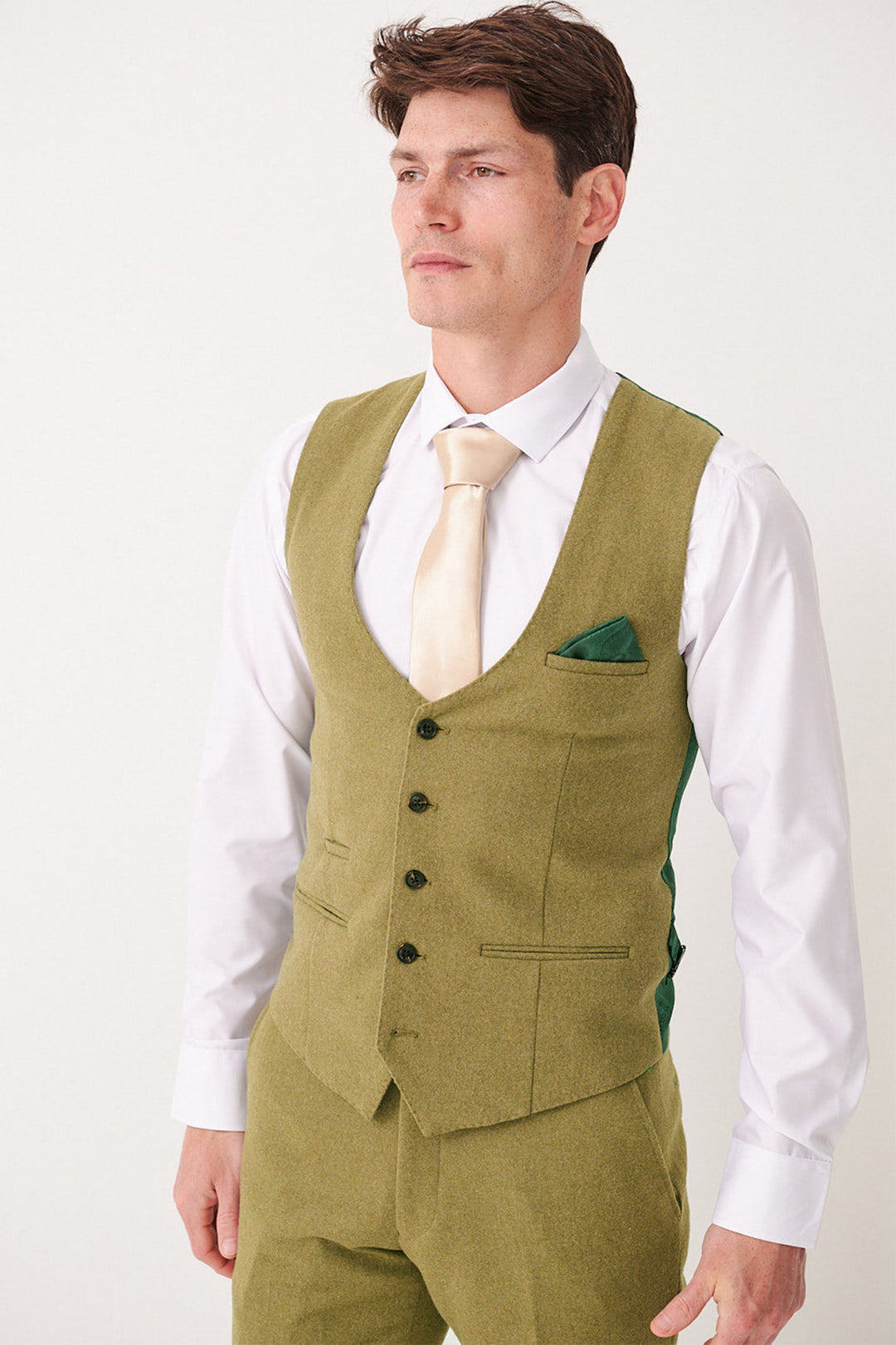 A man wears the Marc Darcy ARCHIE - Moss Tweed Waistcoat, paired with an olive green suit, white shirt, cream tie, and green pocket square, standing before a plain light background.