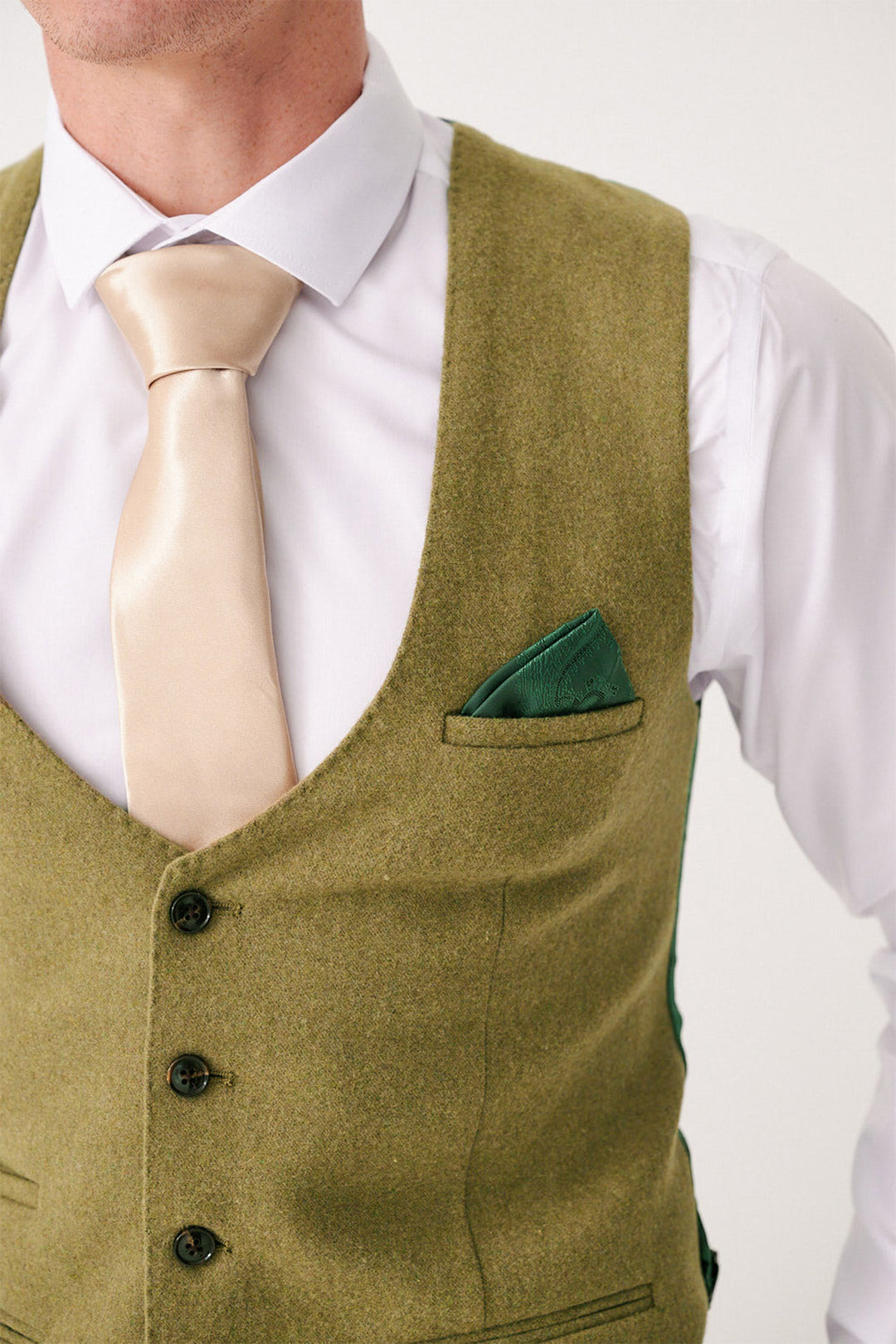 A person in the marcdarcy ARCHIE - Moss Tweed Three Piece Suit, featuring a white dress shirt, light gold satin tie, olive green vest with black buttons and green pocket square. The slim fit suit is shown with the face partially out of frame.