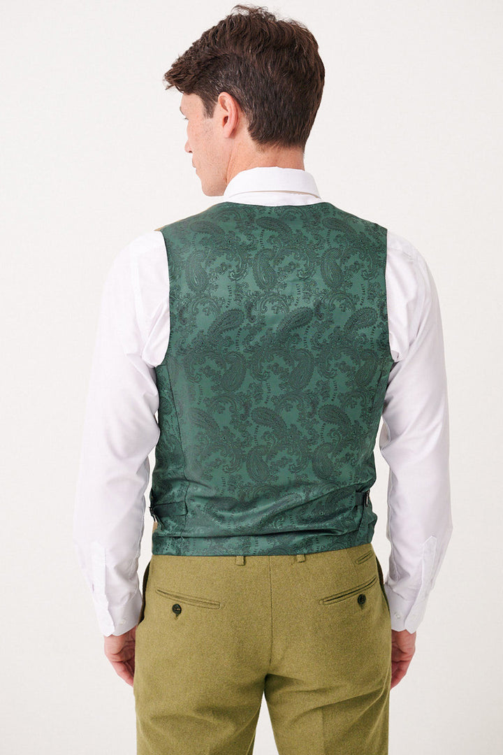 A man stands against a plain light background, wearing a white shirt, olive green trousers, and the ARCHIE - Moss Tweed Waistcoat by Marc Darcy in a green Prince of Wales check pattern.