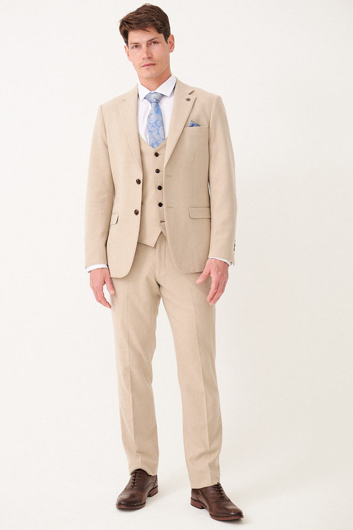 A man stands against a plain background wearing the marcdarcy ARCHIE Stone Tweed Three Piece Suit with a white shirt, blue patterned tie, pocket square, and brown dress shoes.