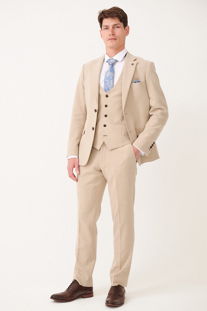 A man with short brown hair and a neutral expression showcases classic men's style in the marcdarcy ARCHIE Stone Tweed Three Piece Suit, paired with a light blue patterned tie and brown dress shoes against a plain background.