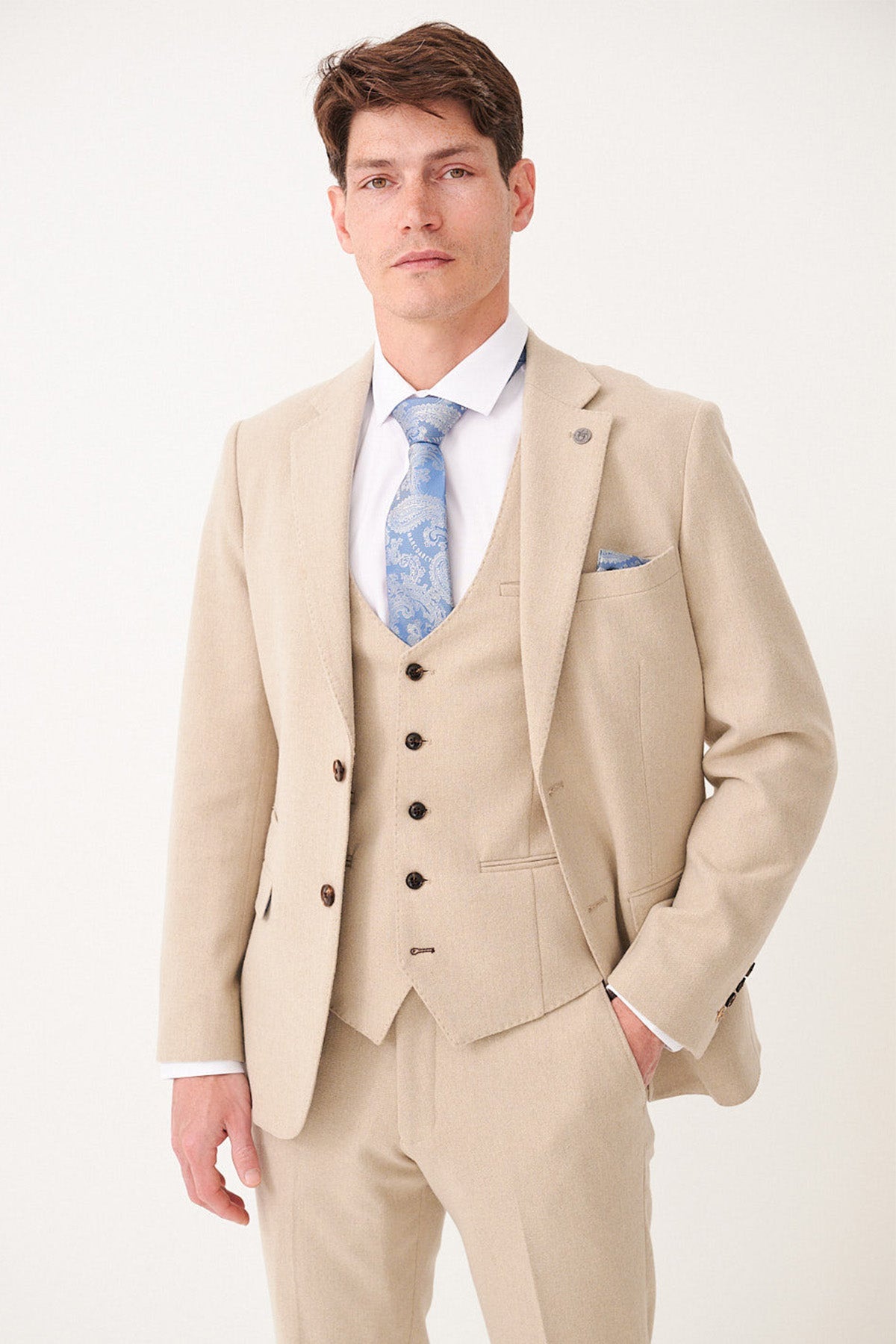 A man wearing the marcdarcy ARCHIE Stone Tweed Blazer stands against a plain light background, styled with a blue patterned tie and pocket square, looking at the camera with a neutral expression.