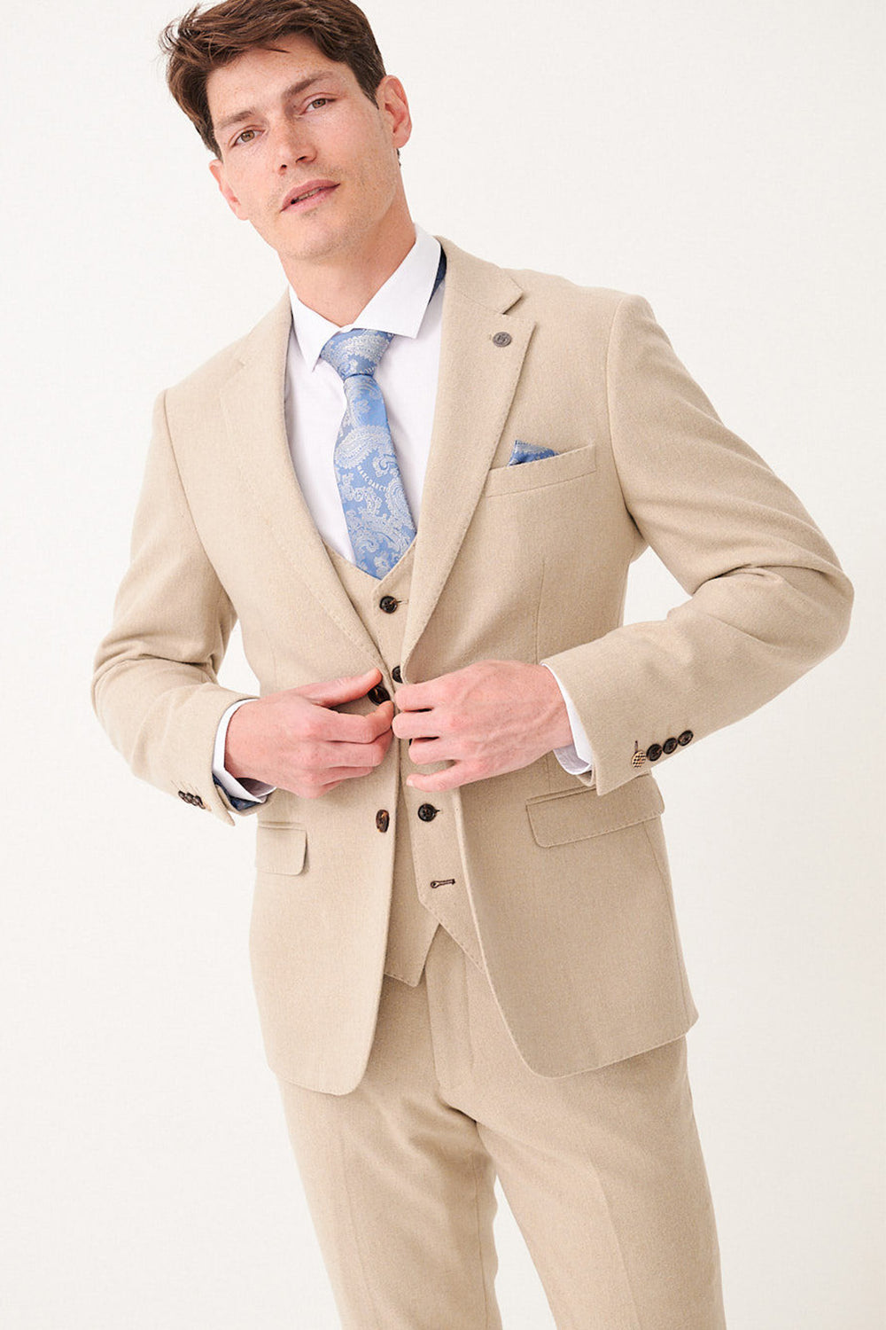 A man wearing the marcdarcy ARCHIE Stone Tweed Blazer, paired with a white shirt and blue patterned tie, stands against a plain white background adjusting the buttons on his slim fit blazer.