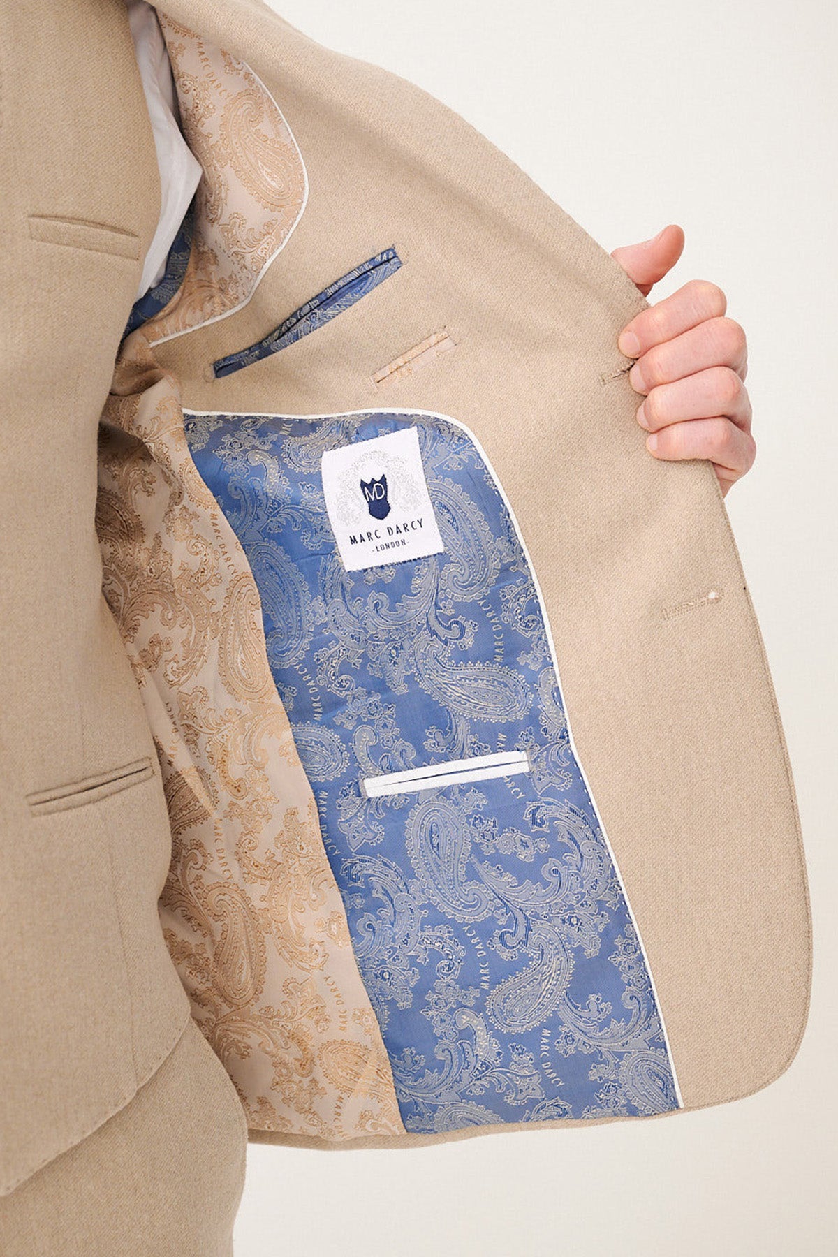 A person opens a marcdarcy ARCHIE - Stone Tweed Blazer to show its inner lining, which features an intricate gold and blue paisley pattern with a white marcdarcy label on the blue section.