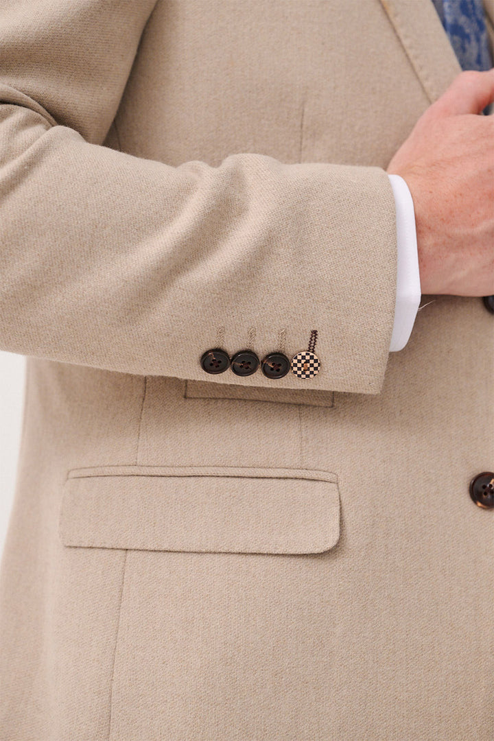 A close-up of a person wearing the marcdarcy ARCHIE Stone Tweed Blazer with a white shirt, showing four brown sleeve buttons and an embroidered patch above the pocket, while adjusting the jacket with their right hand.
