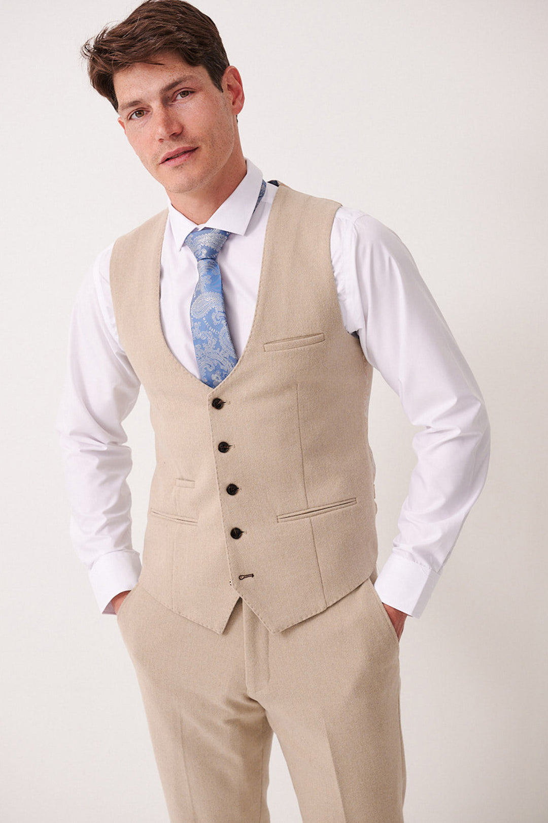 A man in the Marc Darcy ARCHIE - Stone Tweed Waistcoat and matching trousers, paired with a white shirt and light blue patterned tie, stands against a plain backdrop with hands in his pockets.