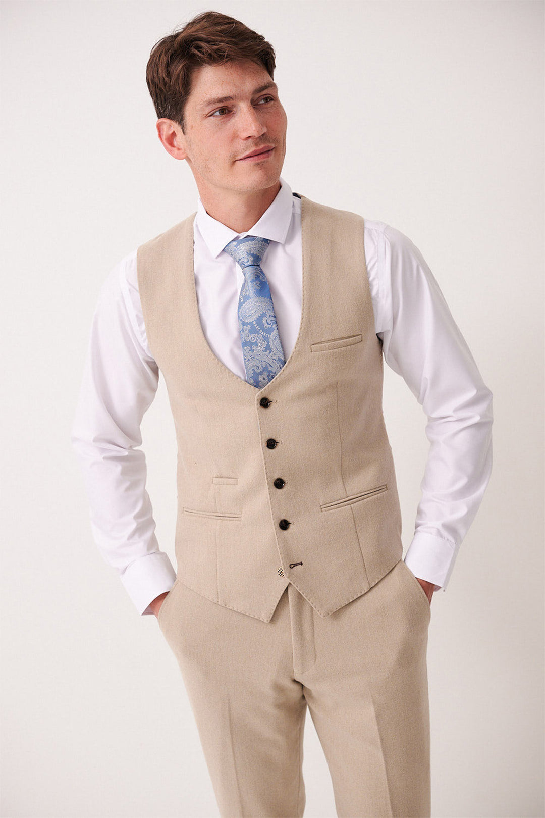 A man wearing the Marc Darcy ARCHIE - Stone Tweed Waistcoat with matching trousers, a white shirt, and a light blue patterned tie stands with his hands in his pockets and smiles slightly against a plain background.