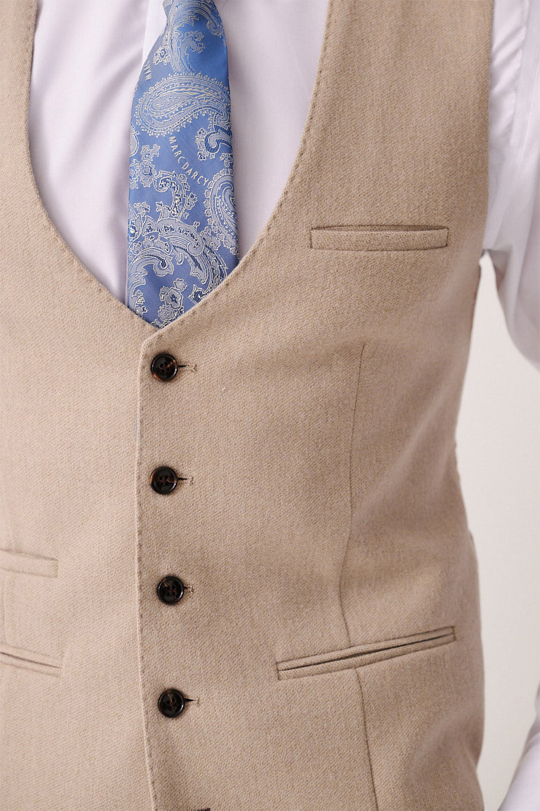 A close-up of a person wearing the Marc Darcy ARCHIE - Stone Tweed Waistcoat over a white shirt with a light blue paisley tie. The waistcoat features dark buttons and three visible pockets.