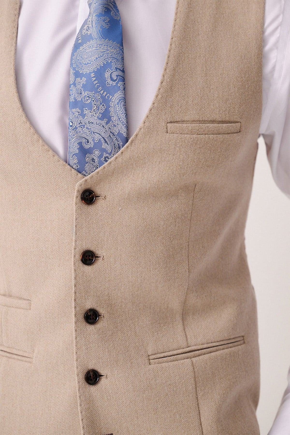 A close-up of a person wearing the Marc Darcy ARCHIE - Stone Tweed Waistcoat over a white shirt with a light blue paisley tie. The waistcoat features dark buttons and three visible pockets.