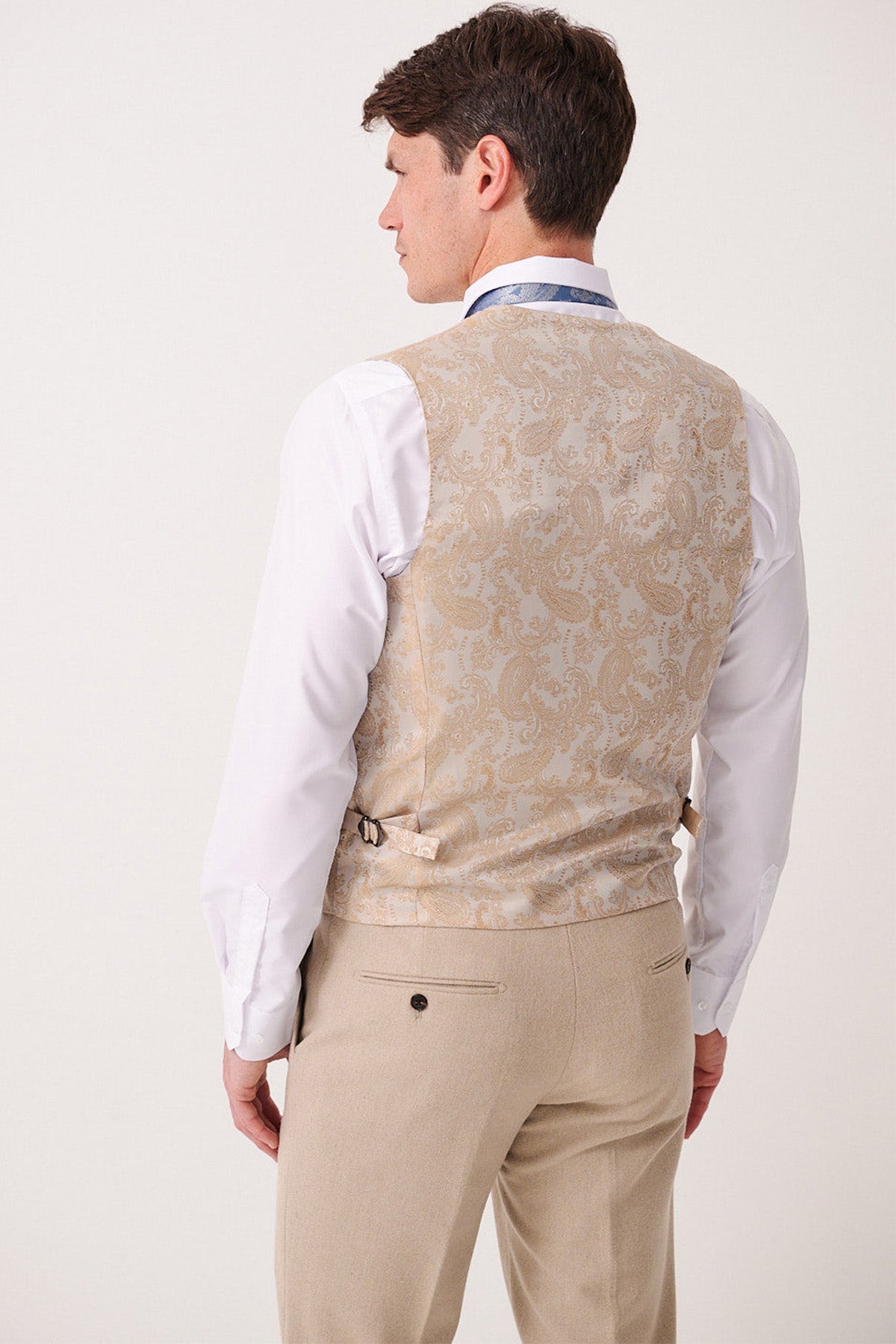 A man is shown from the back wearing the Marc Darcy ARCHIE - Stone Tweed Waistcoat, a slim-fit vest with adjustable side straps, paired with a white shirt and beige trousers. The background is plain and light.