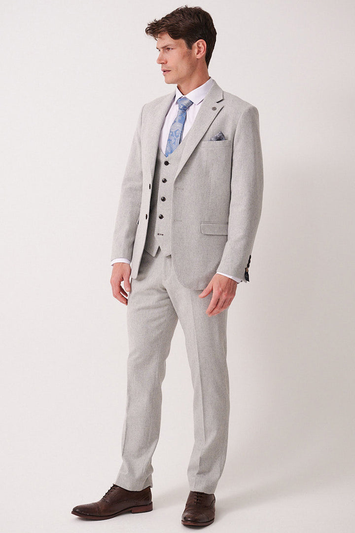 A man wears the marcdarcy ARCHIE Silver Tweed Three Piece Suit with a white shirt, patterned blue tie, dark brown shoes, and a pocket square, standing against a plain light background and looking to the side.