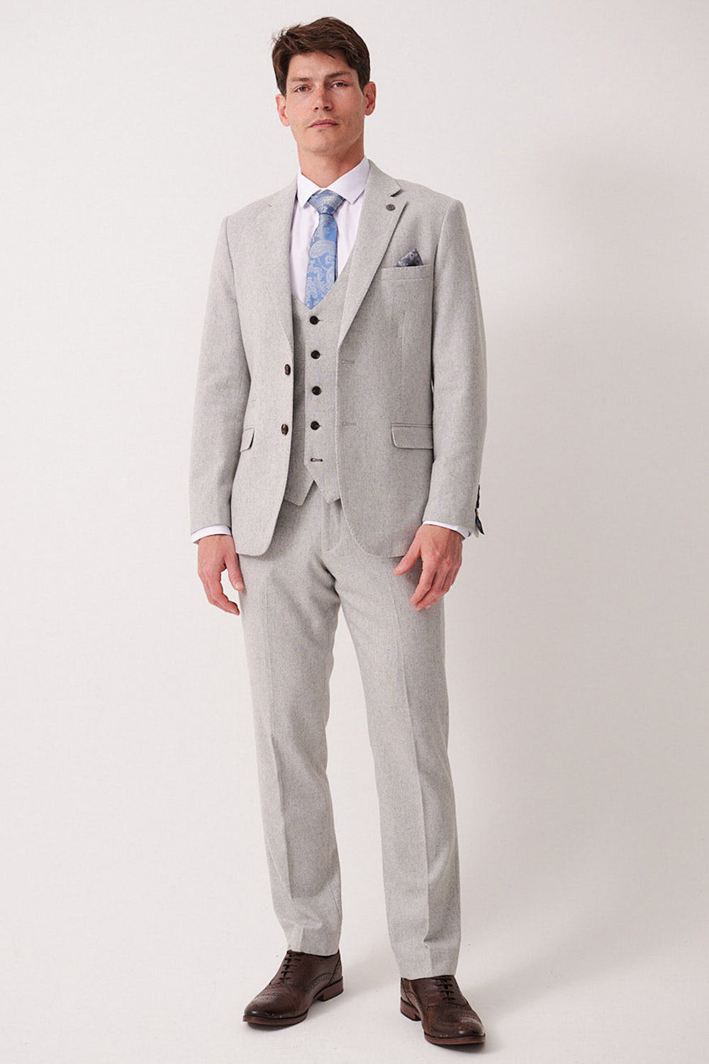 A man stands against a plain background wearing the marcdarcy ARCHIE Silver Tweed Three Piece Suit, paired with a white dress shirt, blue patterned tie, and brown dress shoes. He faces forward with a neutral expression.