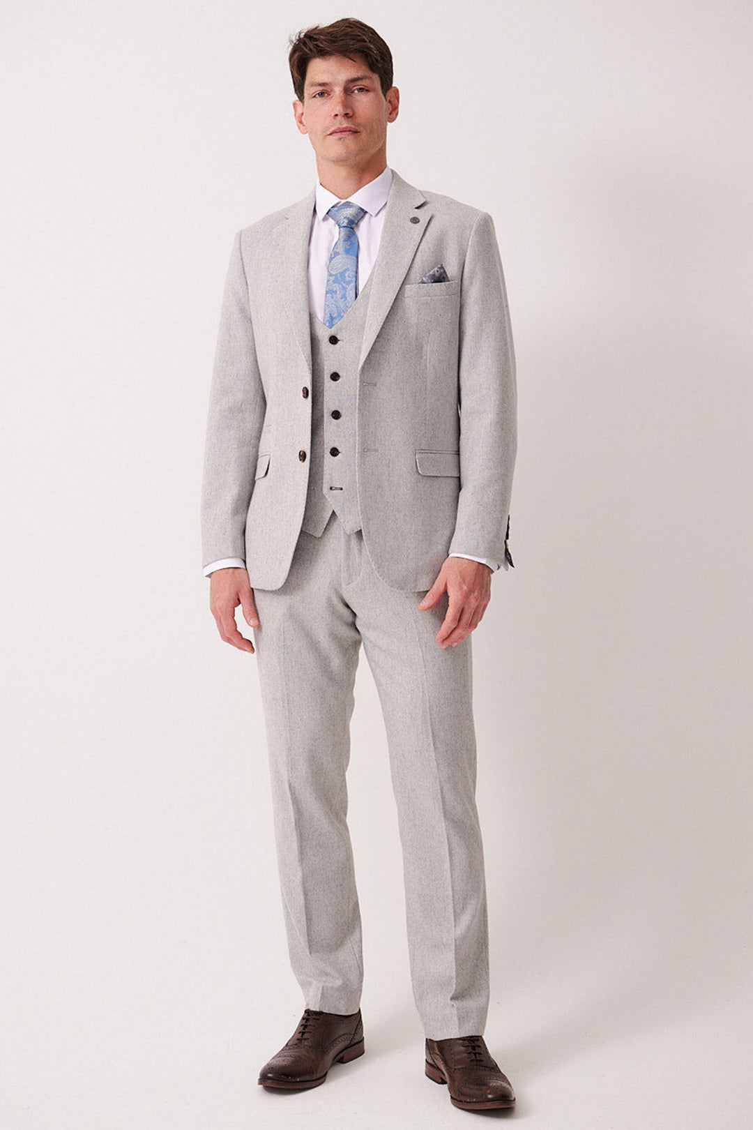 A man poses against a plain light background wearing the marcdarcy ARCHIE Silver Tweed Three Piece Suit, styled with a white shirt, light blue patterned tie, and brown dress shoes. He has short brown hair and a neutral expression.