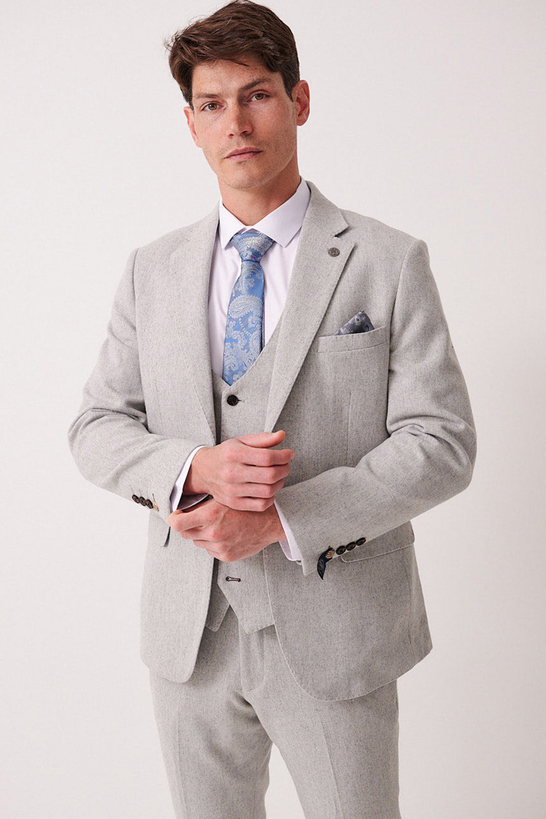A man in a marcdarcy ARCHIE Silver Tweed Blazer, white shirt, light blue paisley tie, and matching pocket square adjusts his jacket and looks confidently at the camera against a plain background.