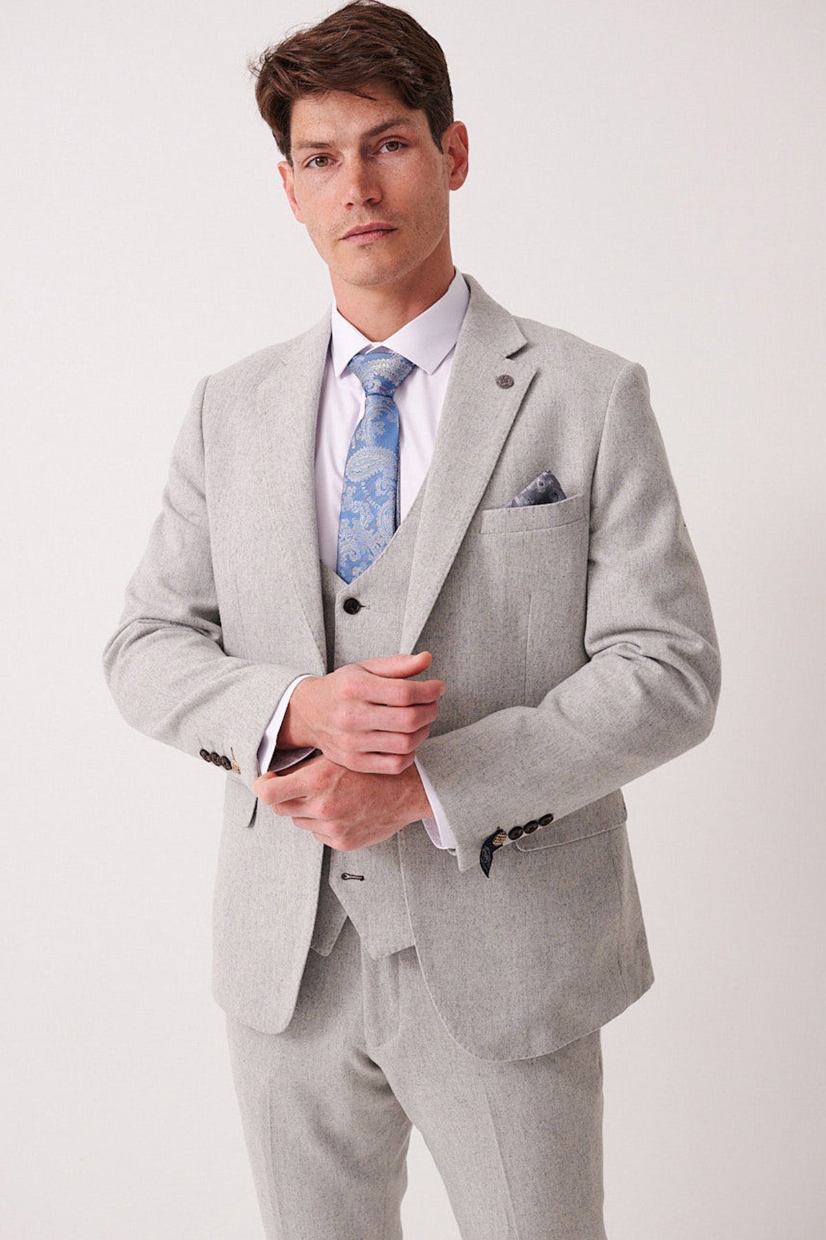 A man in a marcdarcy ARCHIE Silver Tweed Blazer, white shirt, light blue paisley tie, and matching pocket square adjusts his jacket and looks confidently at the camera against a plain background.
