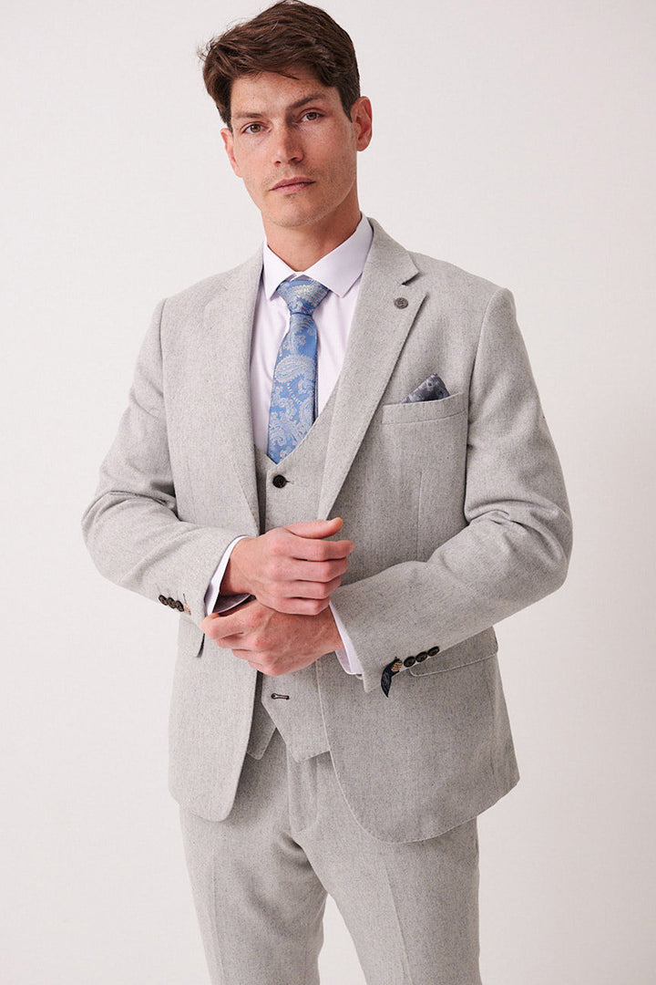 A man in a marcdarcy ARCHIE Silver Tweed Blazer, white shirt, light blue paisley tie, and matching pocket square adjusts his jacket and looks confidently at the camera against a plain background.