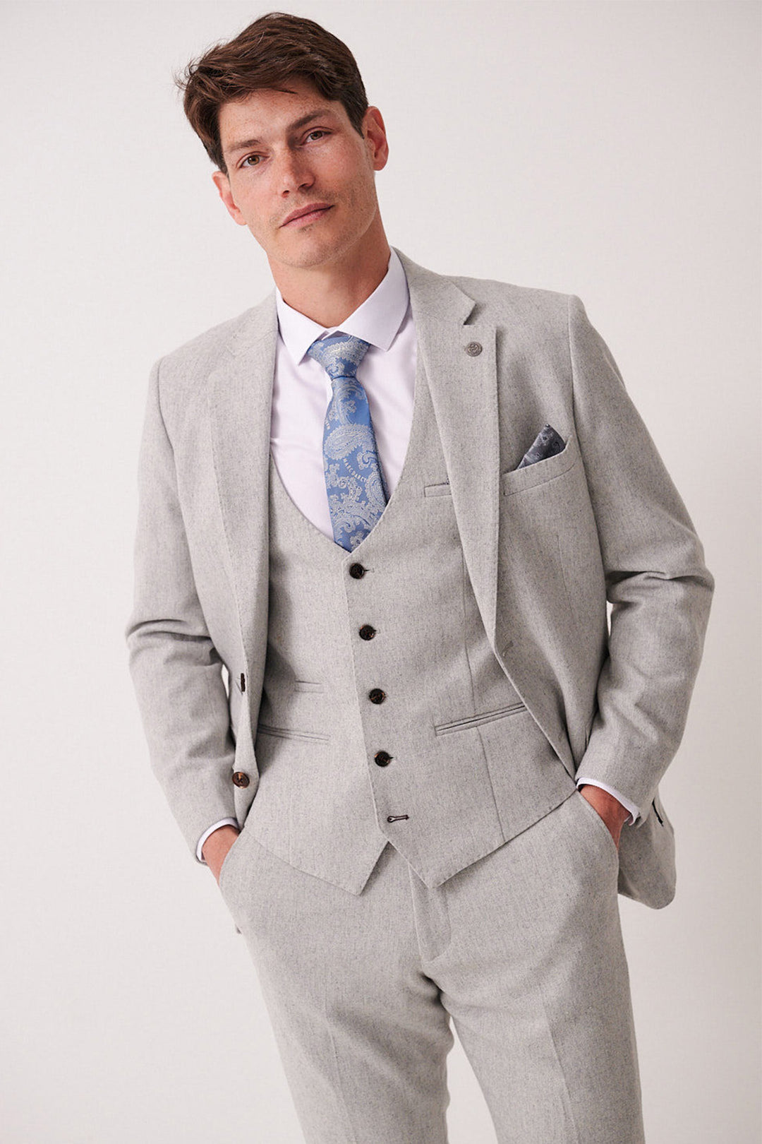 A man wearing a marcdarcy ARCHIE Silver Tweed Blazer with a white shirt, blue patterned tie, and pocket square stands with hands in his pockets, looking at the camera against a plain background.