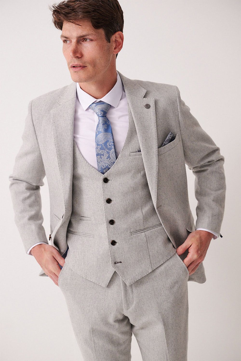 A man stands with his hands on his hips, wearing the marcdarcy ARCHIE Silver Tweed Blazer with a white shirt, blue patterned tie, and matching pocket square, looking to the side against a plain background.