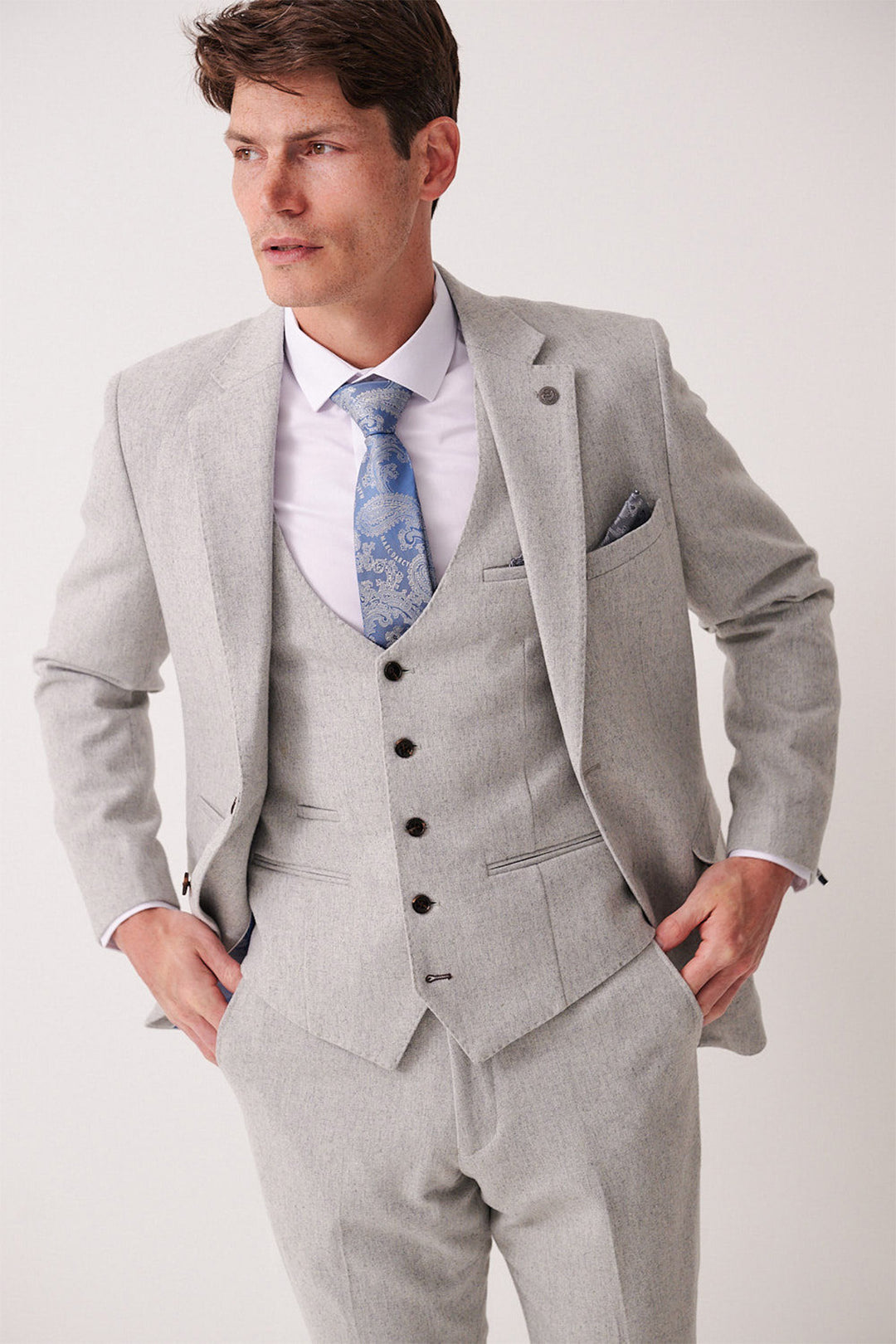A man in the marcdarcy ARCHIE Silver Tweed Three Piece Suit with a matching vest, white shirt, and blue paisley tie stands against a plain light background, hands on his hips.