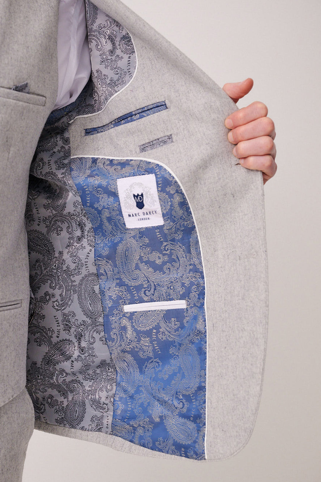 A person holds open the ARCHIE Silver Tweed Three Piece Suit by marcdarcy, showing its slim fit, blue and gray paisley lining, multiple inner pockets, and a label with a black cat logo and "marcdarcy" text.