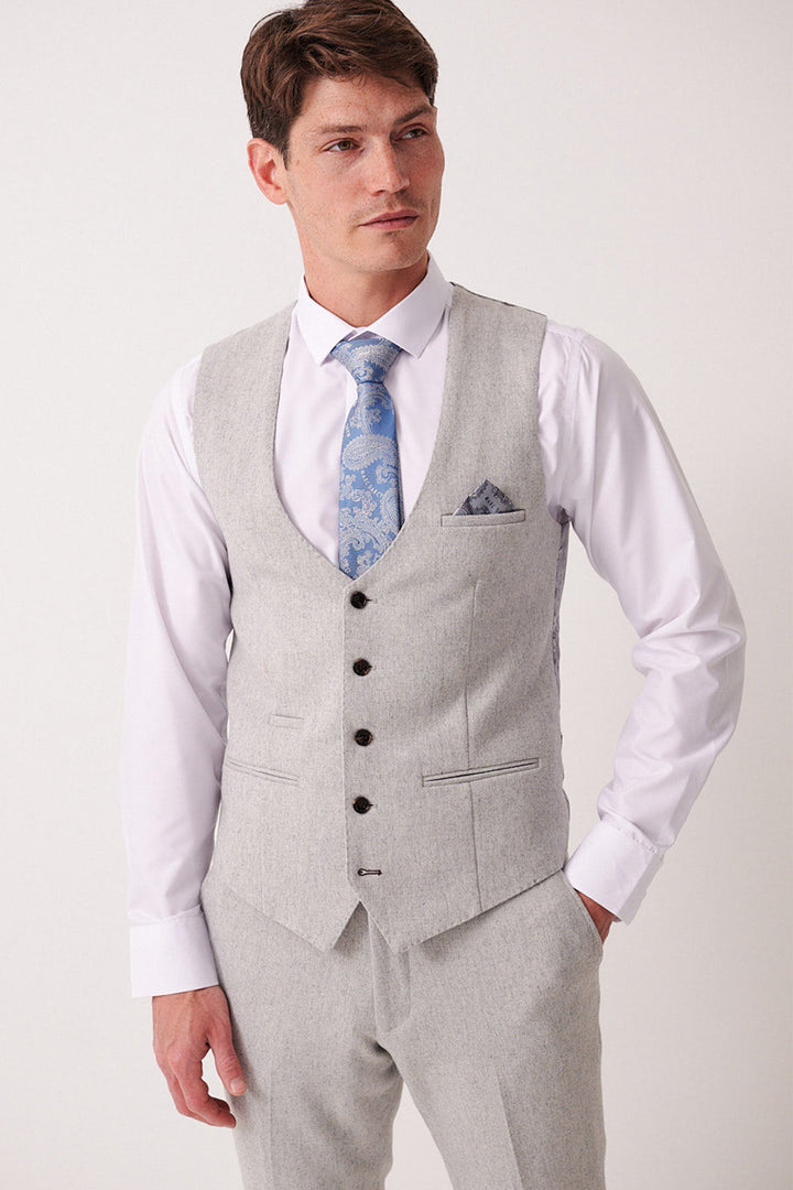 A man stands against a plain light background wearing a Marc Darcy ARCHIE Silver Tweed Waistcoat, matching trousers, a white dress shirt, blue patterned tie, and pocket square.