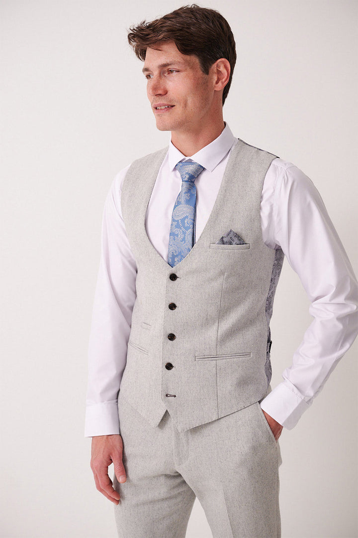 A man wearing the Marc Darcy ARCHIE - Silver Tweed Waistcoat, matching trousers, a white dress shirt, and a blue patterned tie stands slightly turned left, smiling with one hand in his pocket.