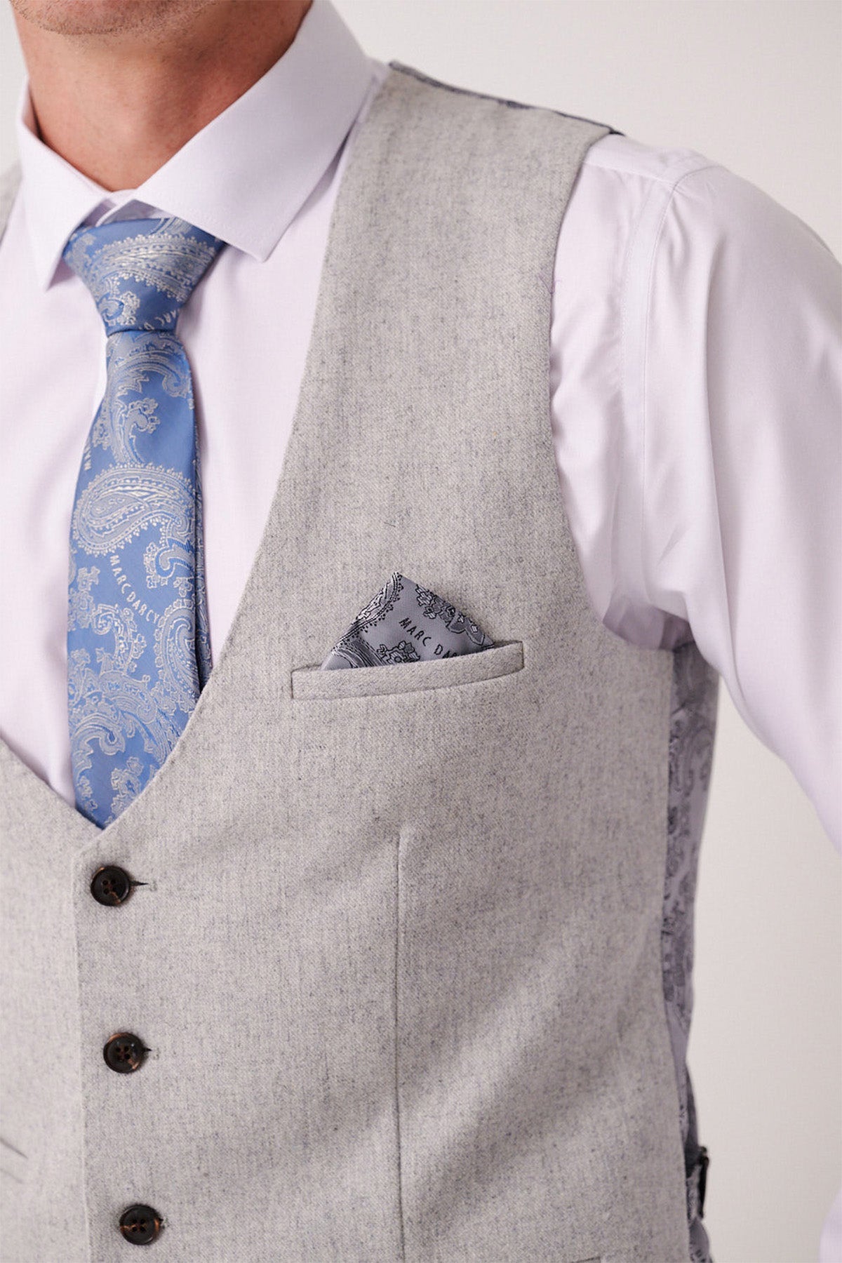 A man wears the Marc Darcy ARCHIE Silver Tweed Waistcoat over a white dress shirt, paired with a blue paisley tie and patterned pocket square. Only his lower face and upper torso are shown.
