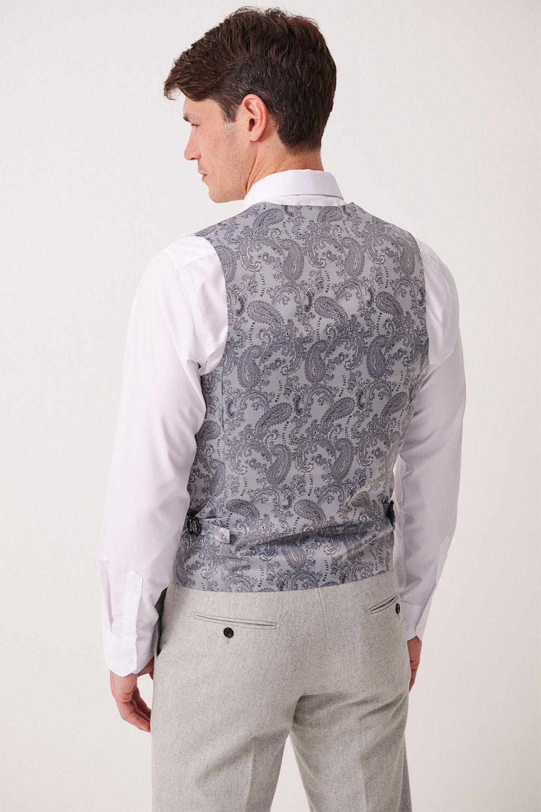 A man faces away, wearing a white dress shirt, light grey trousers, and the Marc Darcy ARCHIE - Silver Tweed Waistcoat featuring an intricate paisley pattern on the back.