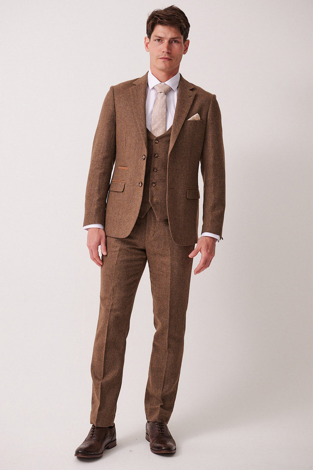 A man stands facing forward in the marcdarcy DEAN Tan Tweed Three Piece Suit, paired with a white shirt, beige tie, and brown shoes, set against a plain light background.