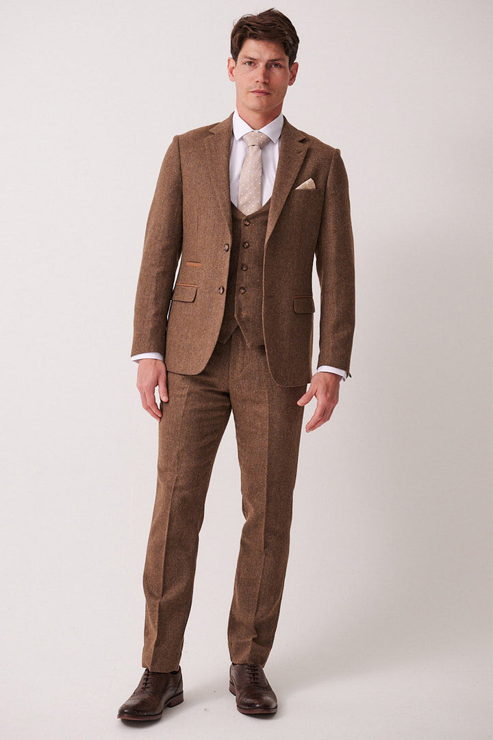 A man stands facing forward in the marcdarcy DEAN Tan Tweed Three Piece Suit, paired with a white shirt, beige tie, and brown shoes, set against a plain light background.
