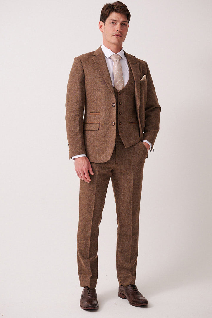 A man stands against a plain background wearing the marcdarcy DEAN Tan Tweed Three Piece Suit with a white shirt, light tie, and brown dress shoes. He has one hand in his pocket and a neutral expression.