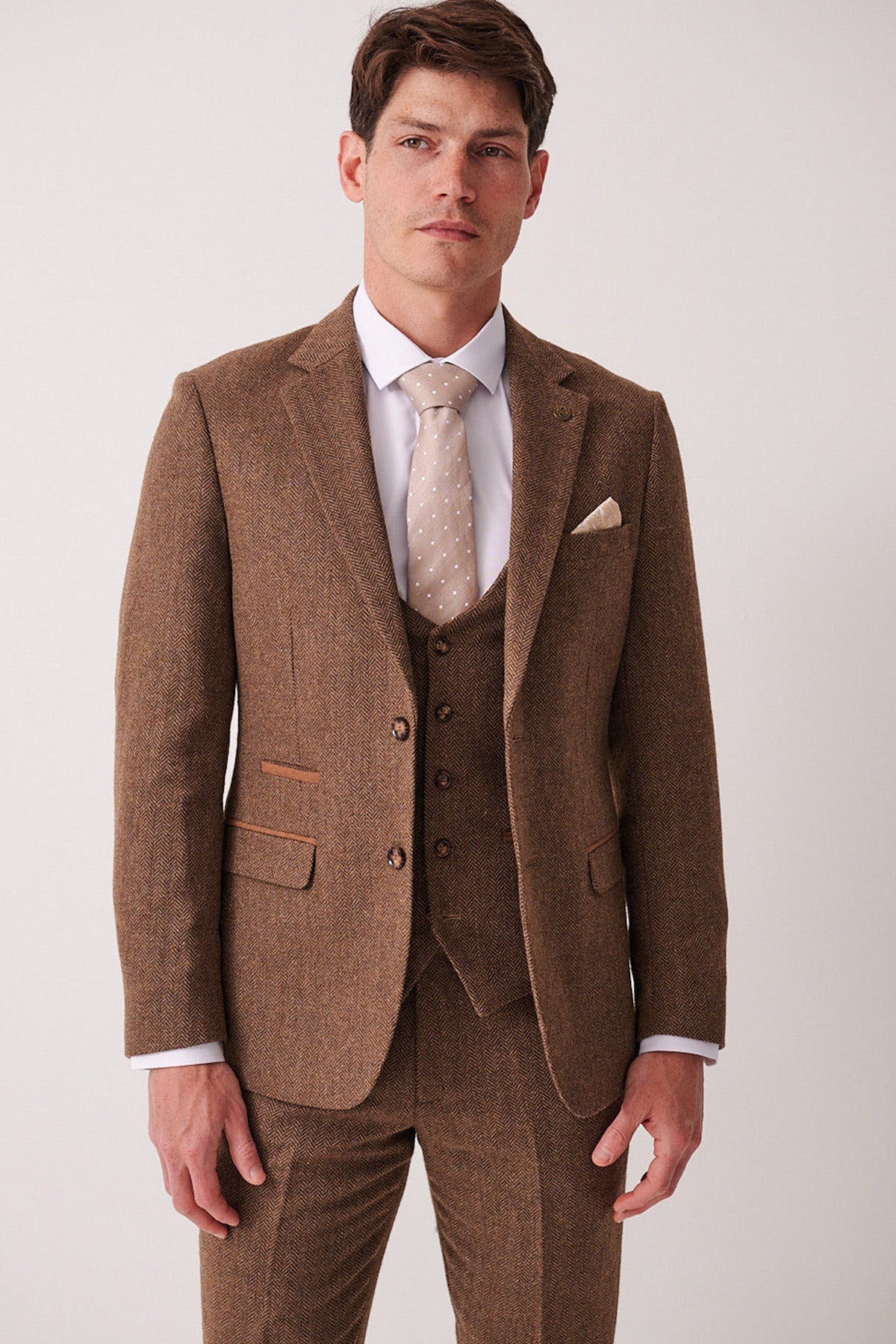 A man wears the marcdarcy DEAN Tan Tweed Blazer with a white dress shirt and beige polka dot tie, standing against a plain light background and looking at the camera with a neutral expression.