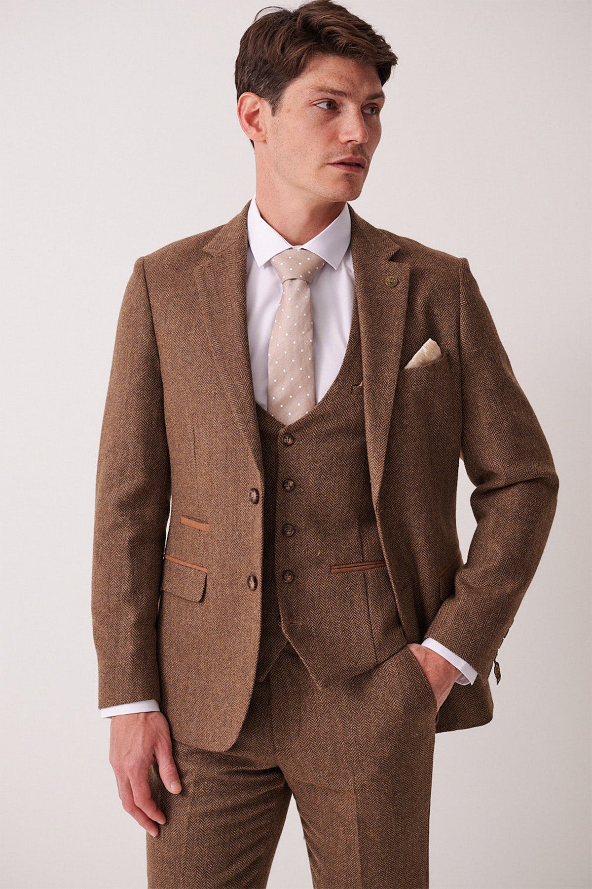 A man in a marcdarcy DEAN Tan Tweed Blazer, paired with a white shirt and light beige polka dot tie, stands with one hand in his pocket, gazing sideways against a plain light background.