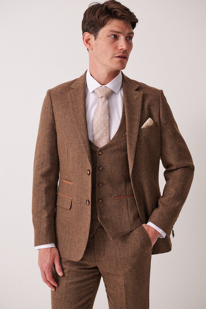 A man in a marcdarcy DEAN Tan Tweed Blazer, paired with a white shirt and light beige polka dot tie, stands with one hand in his pocket, gazing sideways against a plain light background.