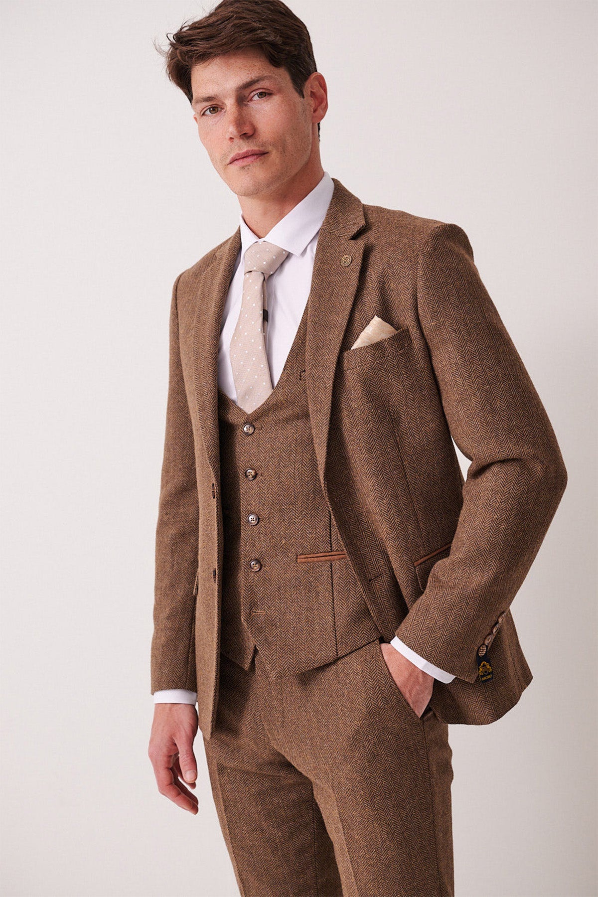 A man wears the marcdarcy DEAN Tan Tweed Blazer with a white shirt, beige tie, and pocket square, standing confidently with one hand in his pocket against a plain light background.